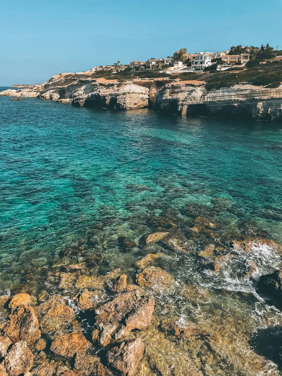 Klares türkisfarbenes Wasser über Felsen an einer Kalksteinküste mit Grotten, oben Häuser auf der Klippe.